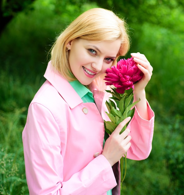 Bella donna con fiore di peonia. Femmina carina nel parco di primavera. Ragazza sorridente con fiore rosa in mano.