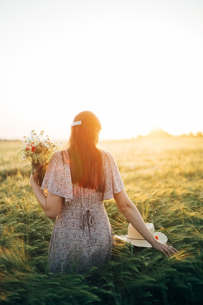 Bella donna con fiori selvatici che si gode il tramonto in un campo d'orzo Momento tranquillo atmosferico Vita lenta rustica Donna elegante che raccoglie fiori e si rilassa la sera in campagna estiva