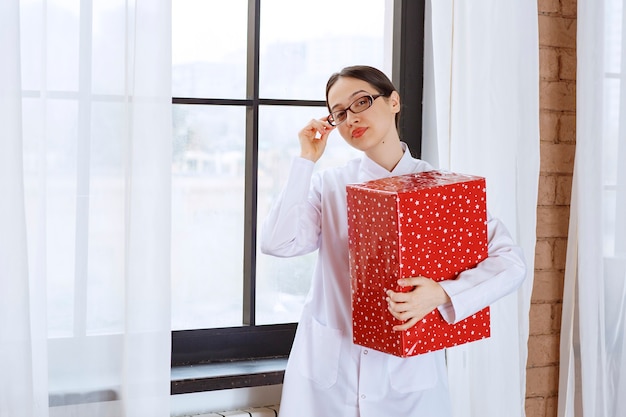 Bella donna con gli occhiali in camice da laboratorio che tiene grande scatola regalo vicino alla finestra.