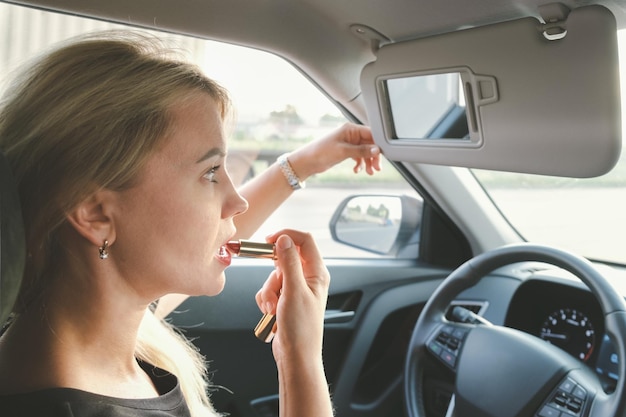 Bella donna con i capelli biondi alla guida di un'auto si dipinge le labbra con il rossetto