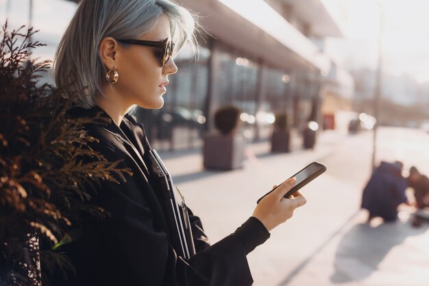 Bella donna con i capelli blu in posa contro il tramonto utilizzando un cellulare e con gli occhiali