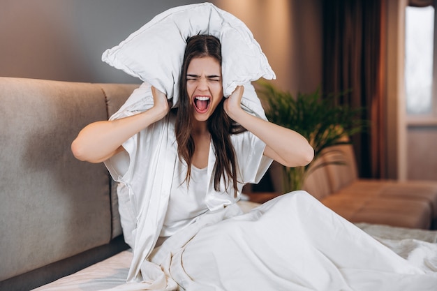 Bella donna con i capelli castani urla mentre è seduta sul letto. La giovane donna si copre le orecchie con un cuscino. Mal di testa da rumore.