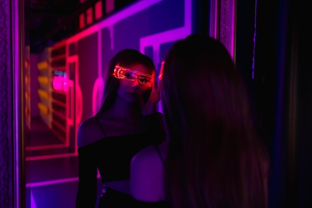 Bella donna con i capelli lunghi in un costume futuristico su sfondo scuro Una ragazza con gli occhiali per realtà virtuale si guarda allo specchio Gioco di realtà aumentata Tecnologia del futuro Concetto di IA Luce al neon BP