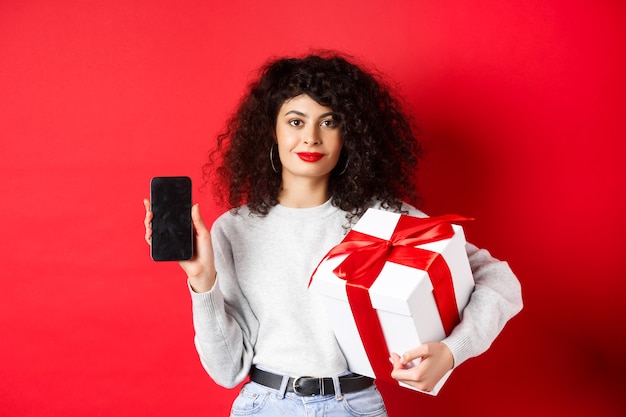 Bella donna con i capelli ricci, mostrando l'app per lo shopping sullo schermo vuoto dello smartphone, tenendo il regalo avvolto in una scatola festiva, in piedi su sfondo rosso.