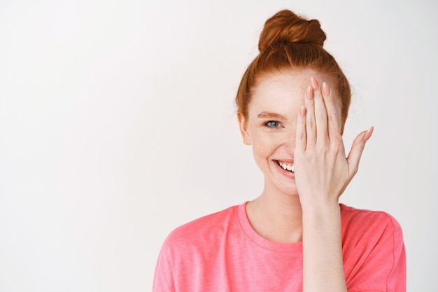 Bella donna con i capelli rossi e gli occhi azzurri che coprono metà del viso, sorridente davanti, in piedi senza trucco sul muro bianco