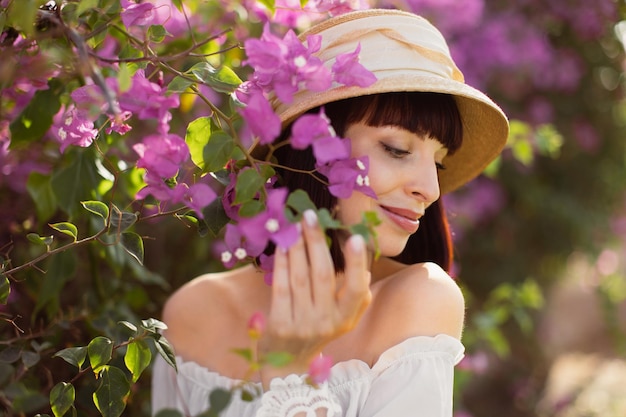 Bella donna con i capelli rossi in abito boho bianco e cappello di paglia vicino all'albero primaverile in fiore