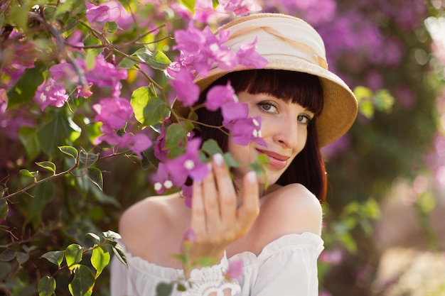 Bella donna con i capelli rossi in abito boho bianco e cappello di paglia vicino all'albero primaverile in fiore