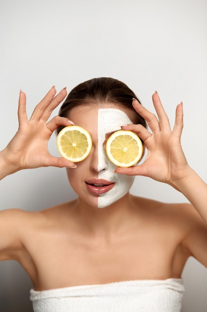 Bella donna con maschera facciale sul viso che tiene fette di limone fresco Bellezza della ragazza Cura della pelle