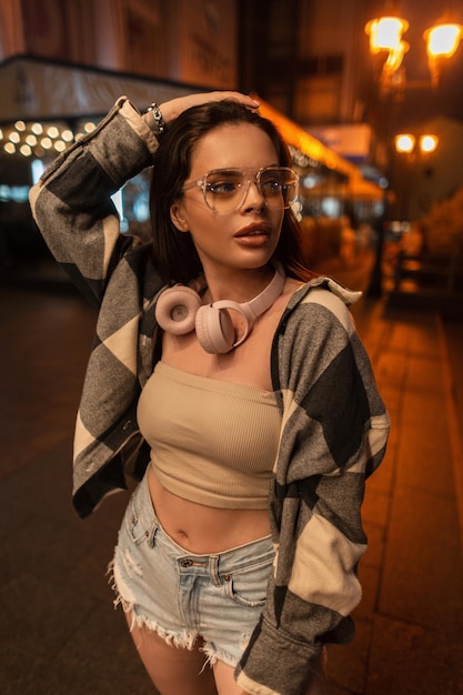 Bella donna con occhiali alla moda e cuffie rosa in una felpa alla moda con un top e pantaloncini di jeans strappati cammina nella città notturna con luce bokeh