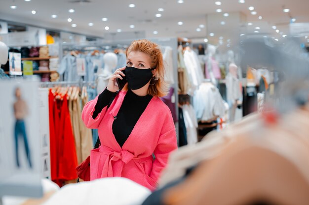 Bella donna con telefono cappotto rosa brillante shopping Mall con maschera protettiva nera