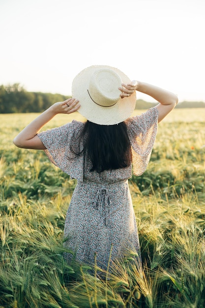 Bella donna con un vestito floreale in piedi in un campo d'orzo alla luce del tramonto Femmina elegante con un cappello di paglia e rilassata nella campagna estiva serale Momento atmosferico vita lenta rustica