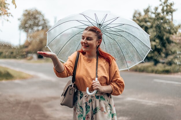 Bella donna con una maschera medica protettiva sta sotto un ombrello trasparente e allunga la mano, catturando gocce d'acqua. Banner orizzontale, copia spazio