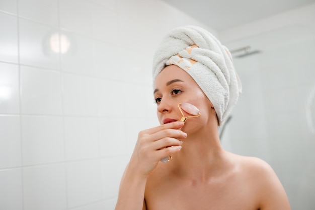 Bella donna con una pelle perfetta che indossa un asciugamano bianco dopo la doccia facendo un massaggio facciale utilizzando un rullo facciale di giada con pietra di quarzo naturale in bagno Concetto di cosmetici naturali levigante delle rughe