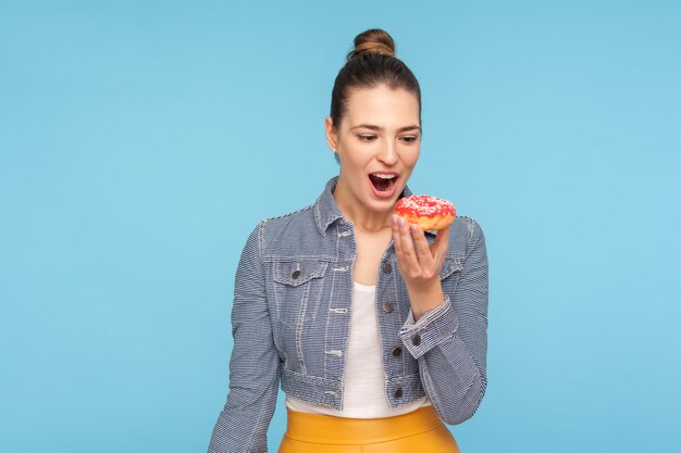 Bella donna contentissima con la crocchia per capelli che guarda una deliziosa ciambella con desiderio, tenendo la bocca aperta e sognando di mordere mangia una ciambella dolce, tentazione di dolciumi zuccherati. ripresa in studio al chiuso