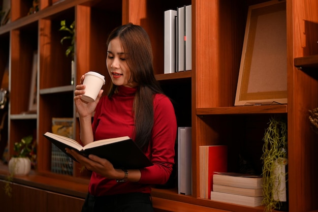 Bella donna d'affari asiatica che sorseggia caffè mentre cerca alcune informazioni sul libro di testo