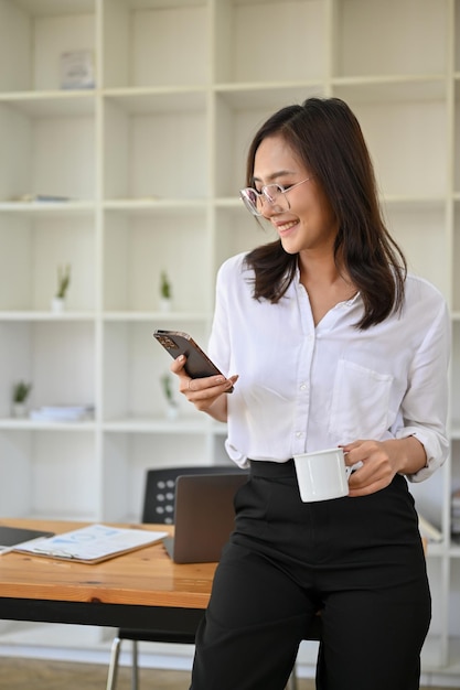 Bella donna d'affari asiatica in piedi nel suo ufficio utilizzando il suo smartphone