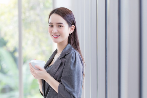 Bella donna d'affari asiatica in piedi tenendo felicemente una tazza di caffè vicino alla finestra sul posto di lavoro