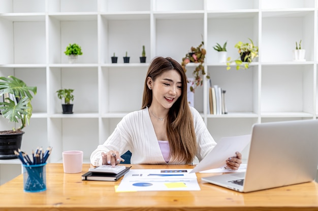 Bella donna d'affari asiatica seduta nel suo ufficio privato, chiacchierando con il suo partner tramite laptop e controllando i documenti, è una dirigente donna di una startup. Concetto di gestione finanziaria.