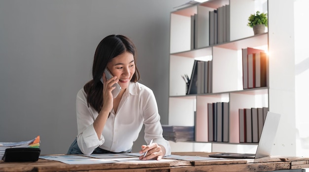 Bella donna d'affari asiatica utilizza lo smartphone per chattare con i clienti al lavoro e riprodurre varie applicazioni di social media con un sorriso rilassato e buon umore