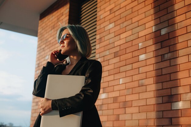 Bella donna d'affari con i capelli blu sta discutendo al telefono mentre posa su un muro di pietra che tiene il suo computer