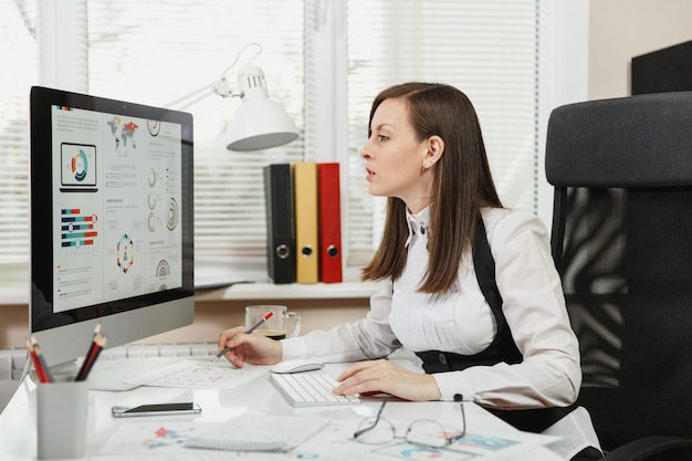 Bella donna d'affari dai capelli castani in giacca e occhiali seduta alla scrivania con una tazza di caffè, lavorando al computer con documenti in ufficio leggero, guardando la telecamera