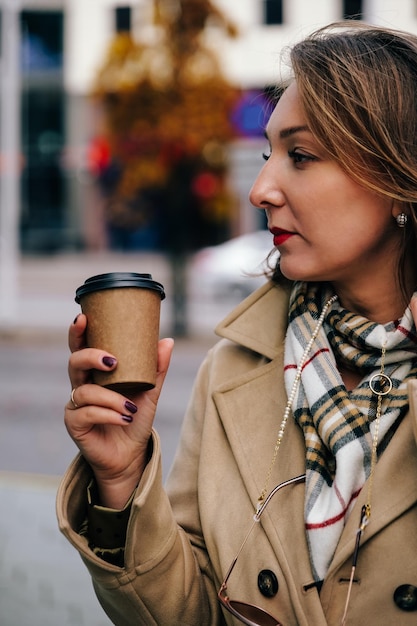 Bella donna d'affari felice che indossa un cappotto beige che cammina in città tenendo il caffè da asporto