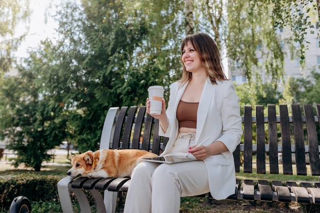 Bella donna d'affari in abito bianco seduto nel parco cittadino, bere caffè e lavorare su tavoletta digitale con il suo cane Welsh Corgi Pembroke