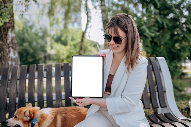 Bella donna d'affari in abito bianco seduto nel parco cittadino con il cane Welsh Corgi Pembroke e dimostra la tavoletta digitale xA