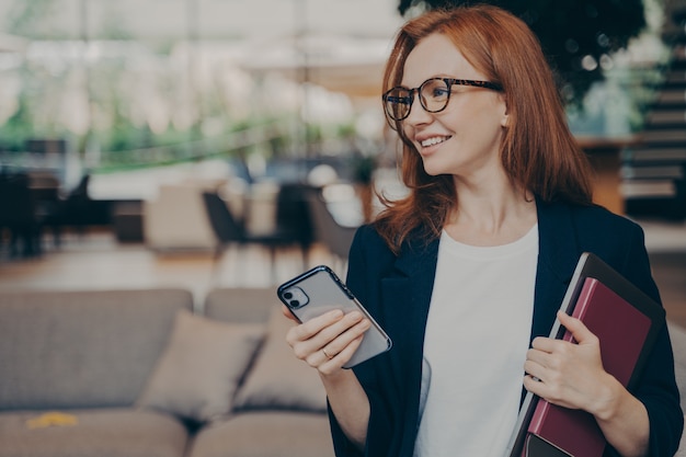 Bella donna d'affari rossa soddisfatta in abito formale che tiene il telefono cellulare in una mano