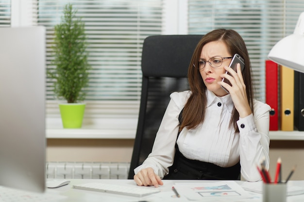 Bella donna d'affari seria in giacca e occhiali seduta alla scrivania, lavorando al computer contemporaneo con documenti in ufficio leggero, parlando al telefono cellulare risolvendo problemi, guardando da parte