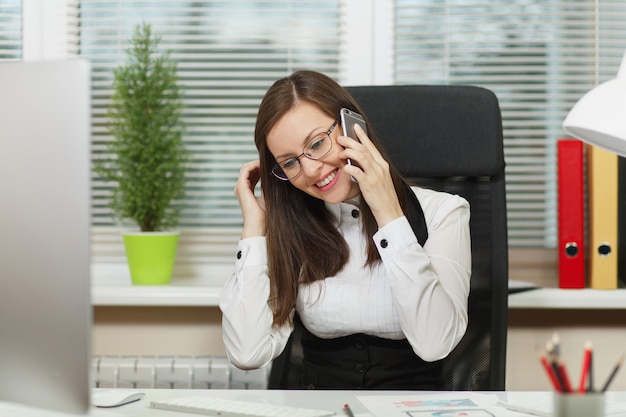 Bella donna d'affari sorridente in giacca e occhiali seduta alla scrivania, lavorando al computer contemporaneo con documenti in ufficio leggero, parlando al cellulare, conducendo una piacevole conversazione