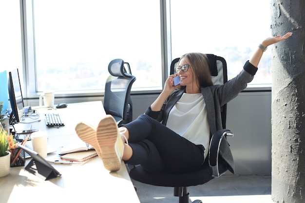 Bella donna d'affari sta parlando al telefono cellulare e sorride mentre è seduta in un ufficio moderno.