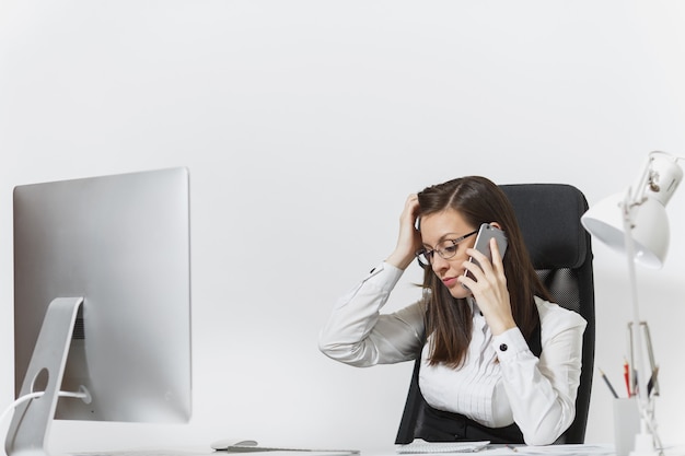 Bella donna d'affari stanca e stressata in tuta seduta alla scrivania, lavorando al computer moderno con documenti in ufficio leggero, parlando al telefono cellulare, risolvendo i problemi