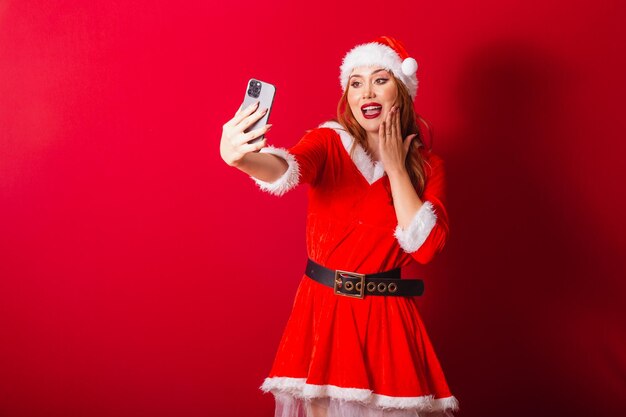 Bella donna dai capelli rossi brasiliana vestita con abiti natalizi Videochiamata di Babbo Natale tramite smartphone