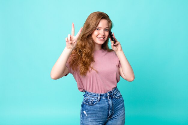 Bella donna dai capelli rossi che sorride e sembra felice, gesticola vittoria o pace e usa uno smartphone