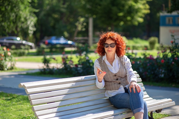 Bella donna dai capelli rossi in estate si siede su una panchina nel parco e utilizza il telefono