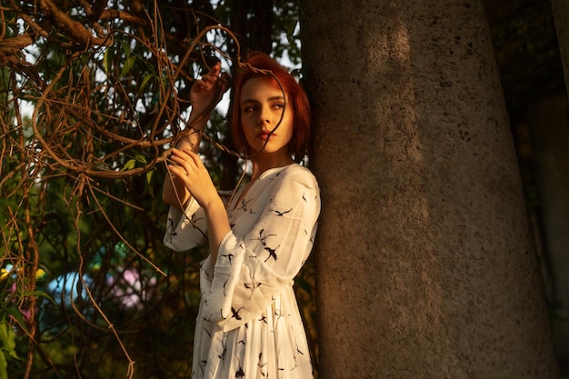 Bella donna dai capelli rossi in un vestito bianco sta posando vicino alla colonna sotto i raggi del sole prima del tramonto