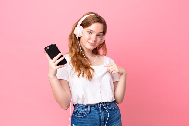 Bella donna dalla testa rossa che sembra arrogante, di successo, positiva e orgogliosa con cuffie e smartphone