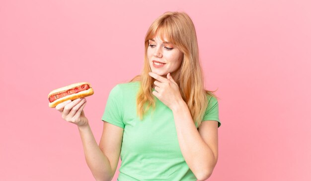 Bella donna dalla testa rossa che sorride con un'espressione felice e sicura con la mano sul mento e tiene un hot dog