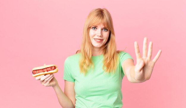 Bella donna dalla testa rossa che sorride e sembra amichevole, mostra il numero quattro e tiene in mano un hot dog