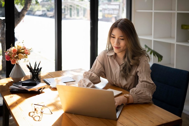 Bella donna di affari asiatica che si siede nell'ufficio moderno luminoso e che utilizza il computer portatile