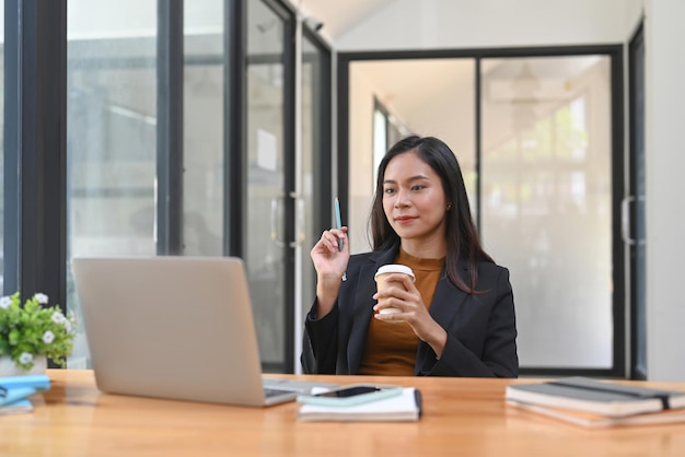 Bella donna di affari che tiene tazza di caffè e utilizza il computer portatile alla scrivania