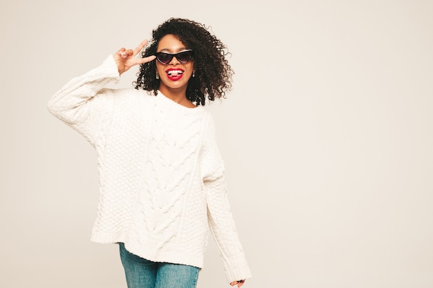 Bella donna di colore con l'acconciatura di riccioli afro e labbra rosse. Modello sorridente in vestiti di jeans alla moda e maglione invernale.