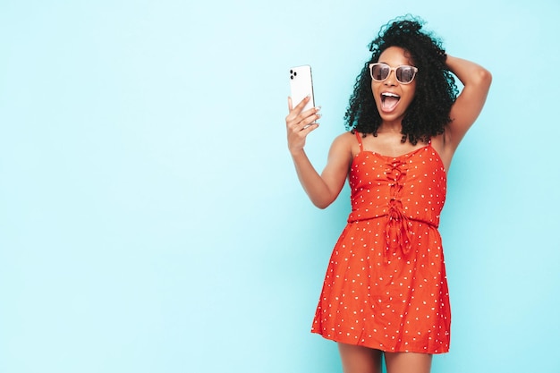 Bella donna di colore con l'acconciatura di riccioli afro Modello sorridente vestito in abito estivo rosso Femmina spensierata sexy in posa vicino al muro blu in studio Selfie abbronzato e allegro che prende