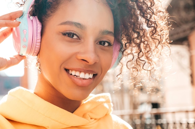 Bella donna di colore con l'acconciatura di riccioli afroModello sorridente in felpa con cappuccio giallaFemmina sexy spensierata che si gode l'ascolto di musica in cuffie wireless Posa su sfondo di strada al tramonto