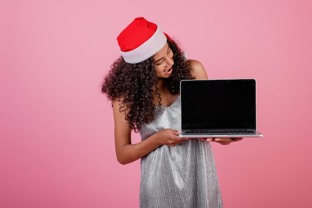 Bella donna di colore con lo schermo del copyspace sul computer portatile che porta il cappello di Santa isolato sopra il rosa