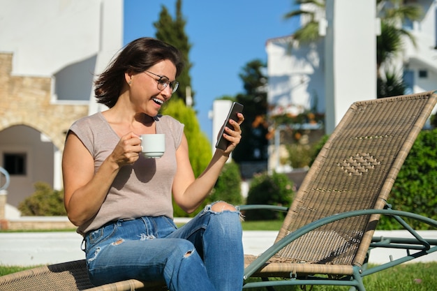 Bella donna di mezza età che riposa leggendo bevendo caffè in chat. Donna seduta sul lettino da giardino sul prato vicino a casa. Stile di vita, tempo libero, tecnologia, bellezza delle persone mature