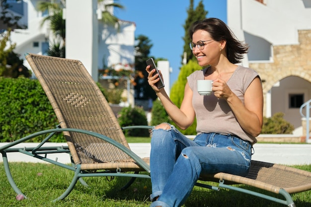 Bella donna di mezza età che riposa seduta sul lettino sul prato verde, leggendo lo smartphone tramite Internet, godendosi una tazza di caffè rilassante in una soleggiata giornata estiva. Vacanze, stile di vita, tempo libero delle persone anziane