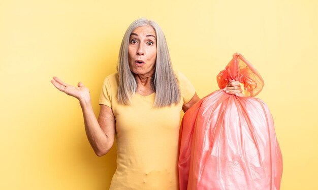 Bella donna di mezza età che sembra sorpresa e scioccata, con la mascella caduta in possesso di un oggetto. sacchetto della spazzatura di plastica