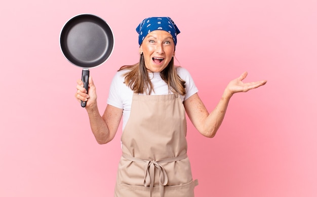 Bella donna di mezza età che si sente felice e stupita per qualcosa di incredibile e tiene in mano una padella. concetto di chef